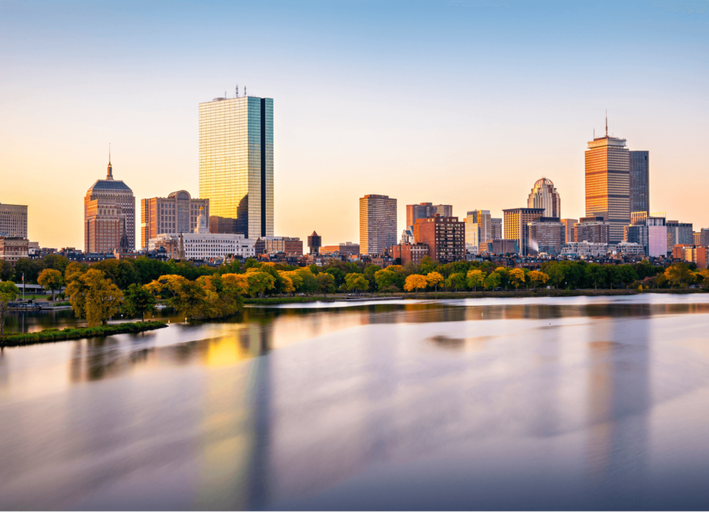 A city skyline at sunset with tall buildings reflecting golden light. The foreground features a calm body of water and lush green trees. The sky is clear with a warm gradient from blue to orange.