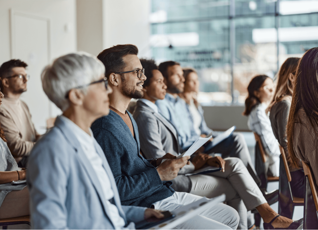 A diverse group of people seated in rows at a conference or seminar, listening attentively. They are casually dressed, holding papers or notepads, with a modern office setting visible in the background.