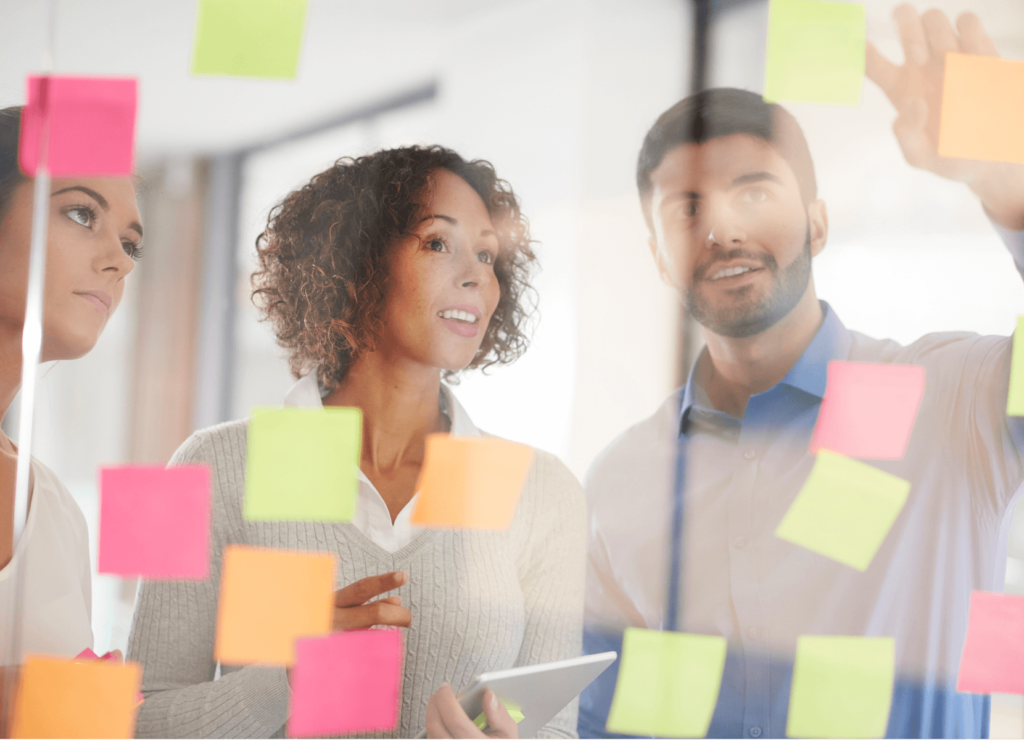 Three people engaged in discussion stand behind a glass wall covered with colorful sticky notes. One holds a tablet, while another points at the notes. The scene suggests brainstorming or planning in an office setting.
