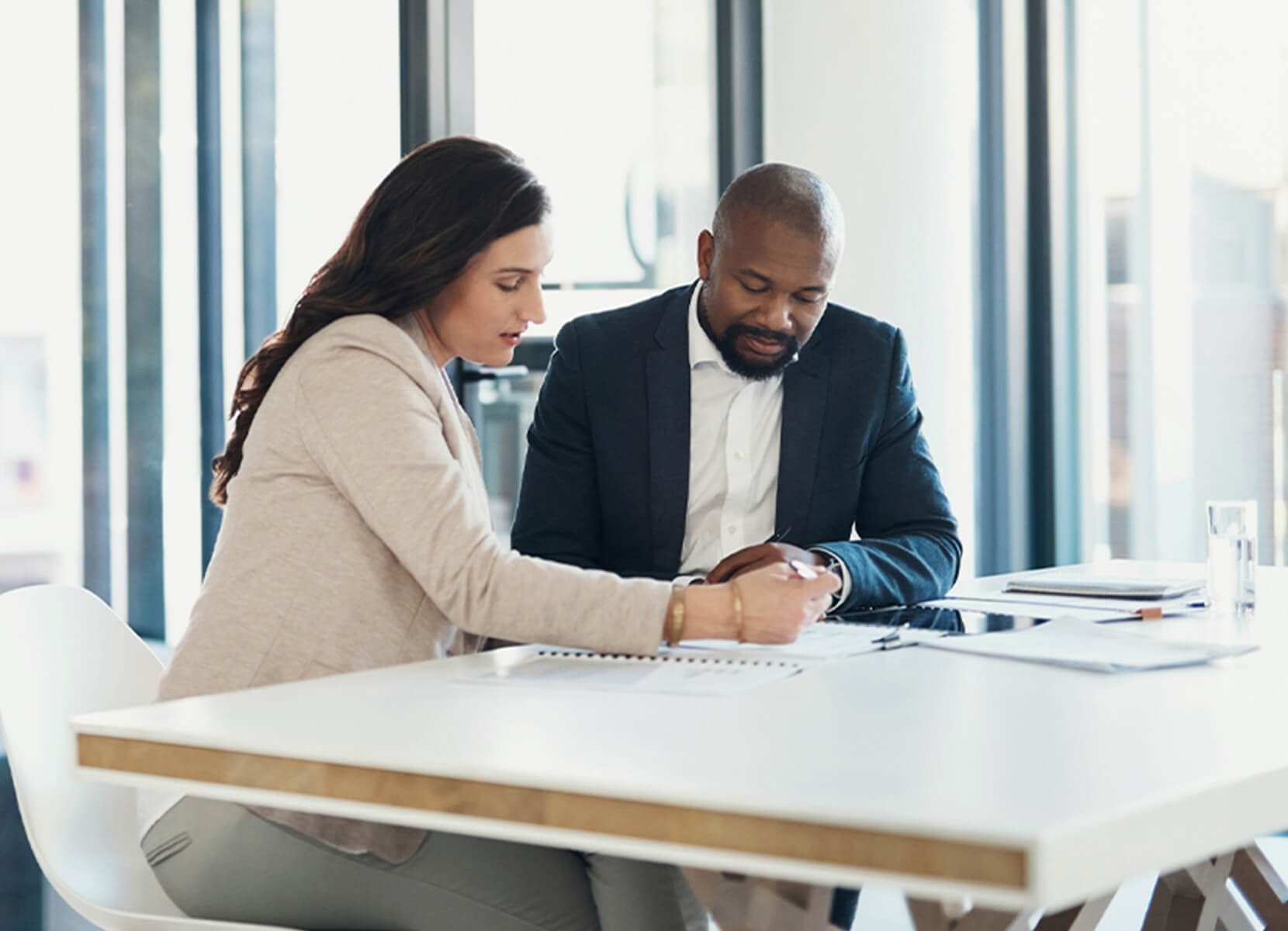 A woman and a man are seated at a table in an office setting. They are reviewing documents together.