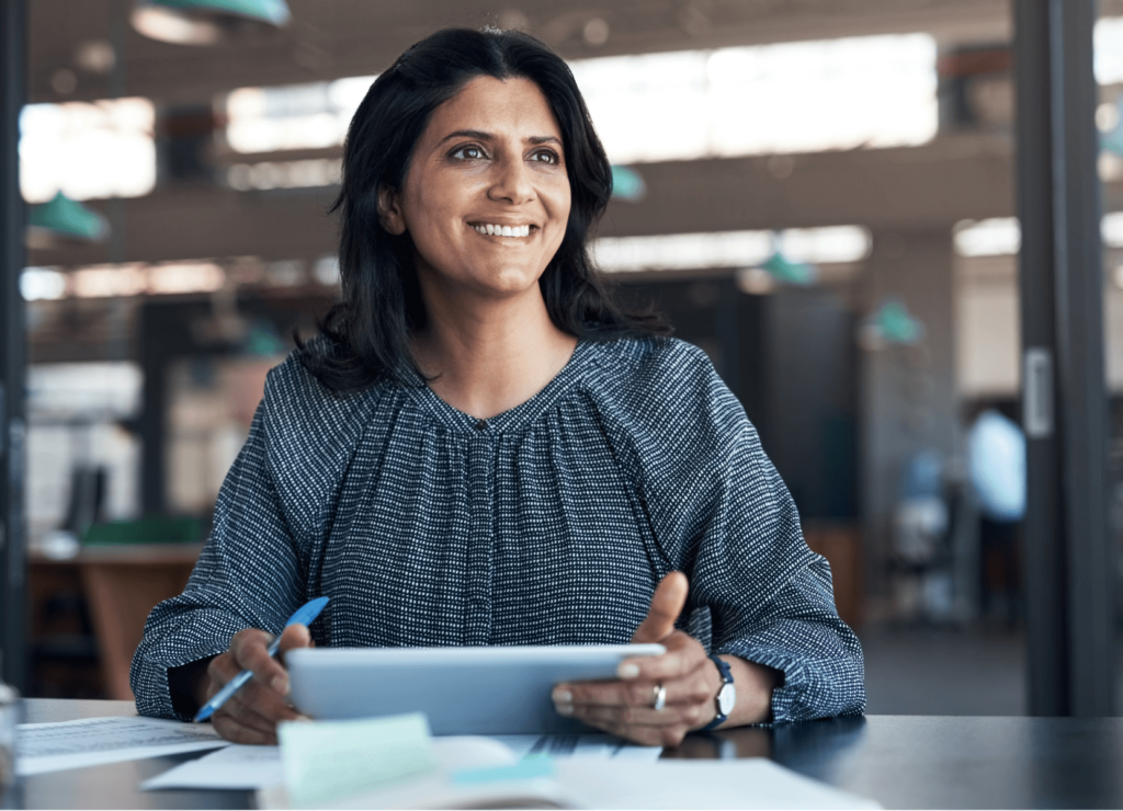 A woman with dark hair smiles while sitting at a desk, holding a tablet. She is wearing a patterned blouse and surrounded by office documents. The office background is softly blurred, suggesting a busy work environment.