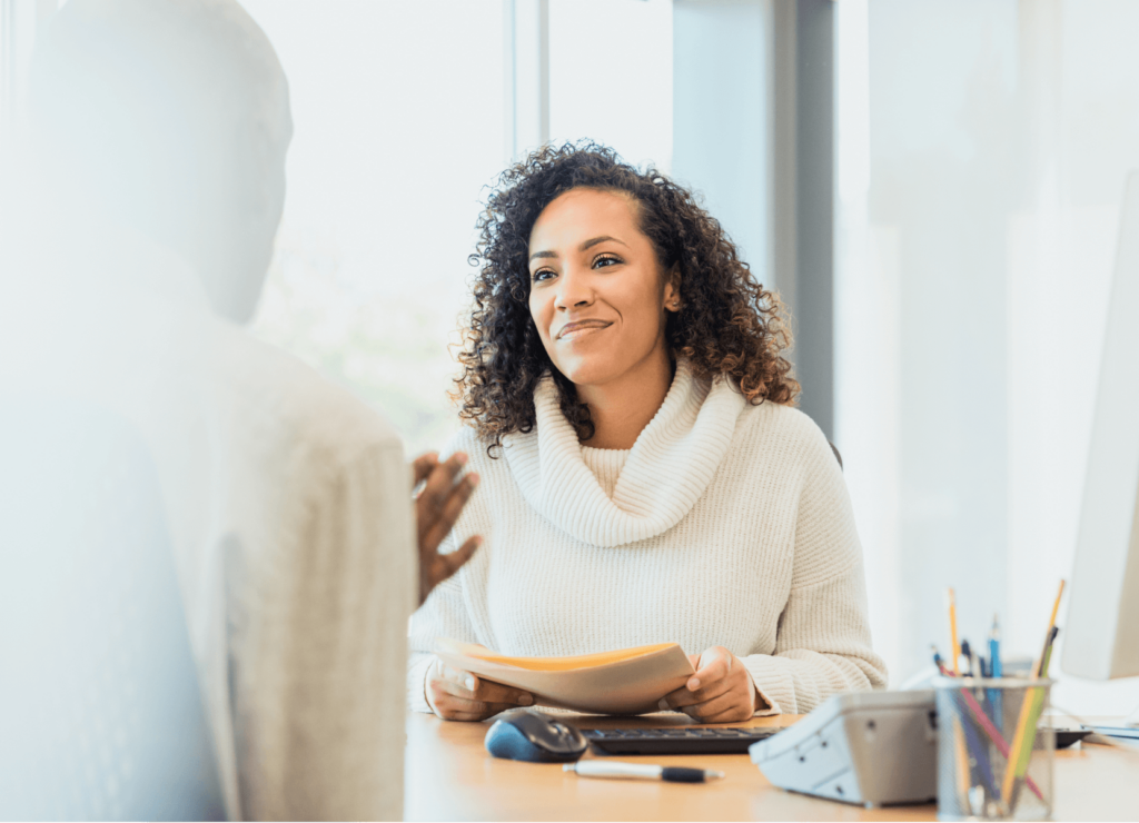 A woman with curly hair, wearing a white sweater, holds documents and smiles while sitting at a desk. She is engaged in a conversation with a person whose back is to the camera. The desk has office supplies, and large windows are in the background.
