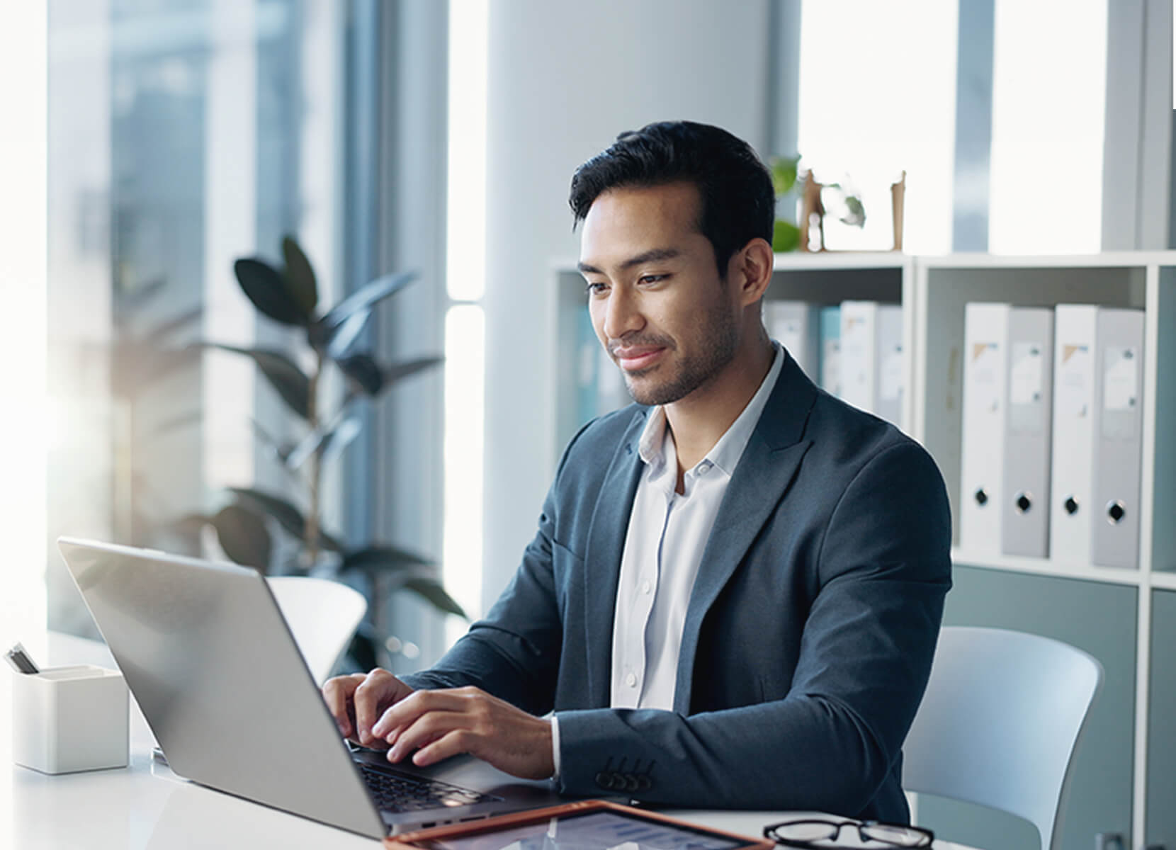 Young man works on a laptop while sitting at a desk in a brightly lit room